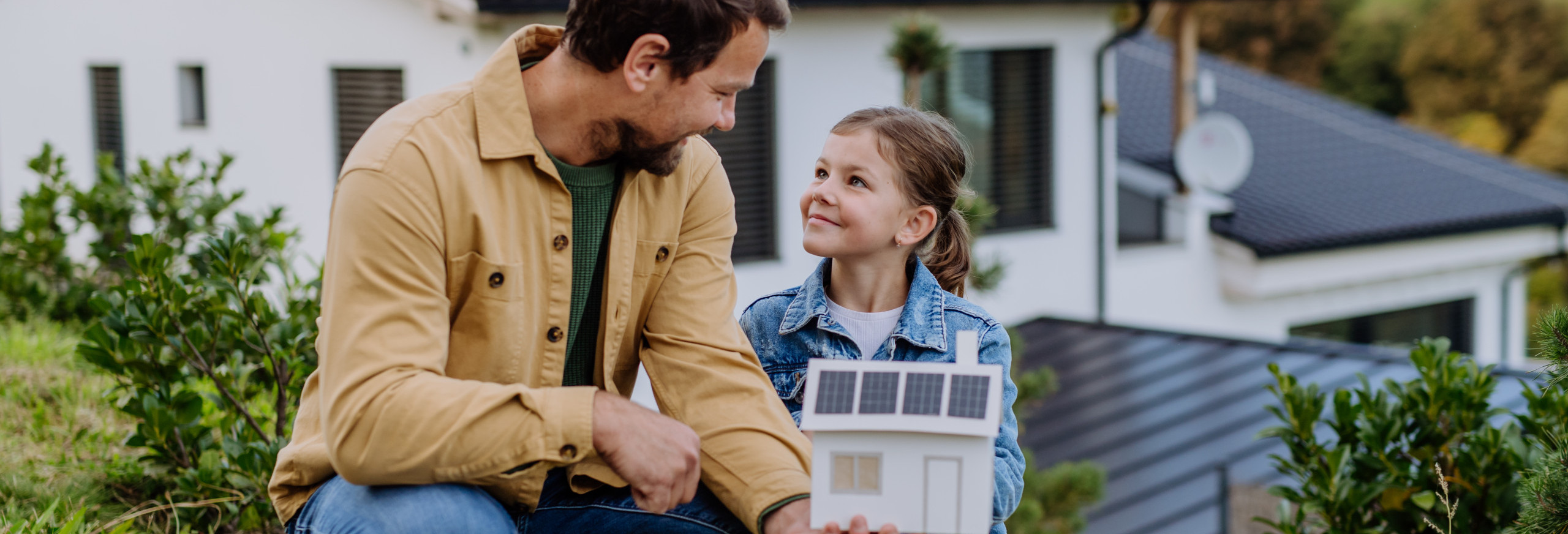 Ein Vater mit Kind sitzen vor einem Haus, auf dessen Dach sich eine Solaranlage befindet.