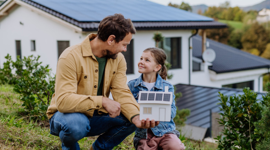 Ein Vater mit Kind sitzen vor einem Haus, auf dessen Dach sich eine Solaranlage befindet.