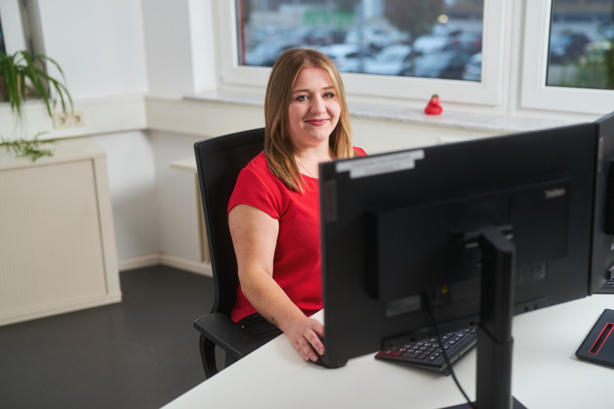 Mitarbeiterin sitzt lächelnd am Arbeitsplatz im Büro und trägt ein rotes Shirt.